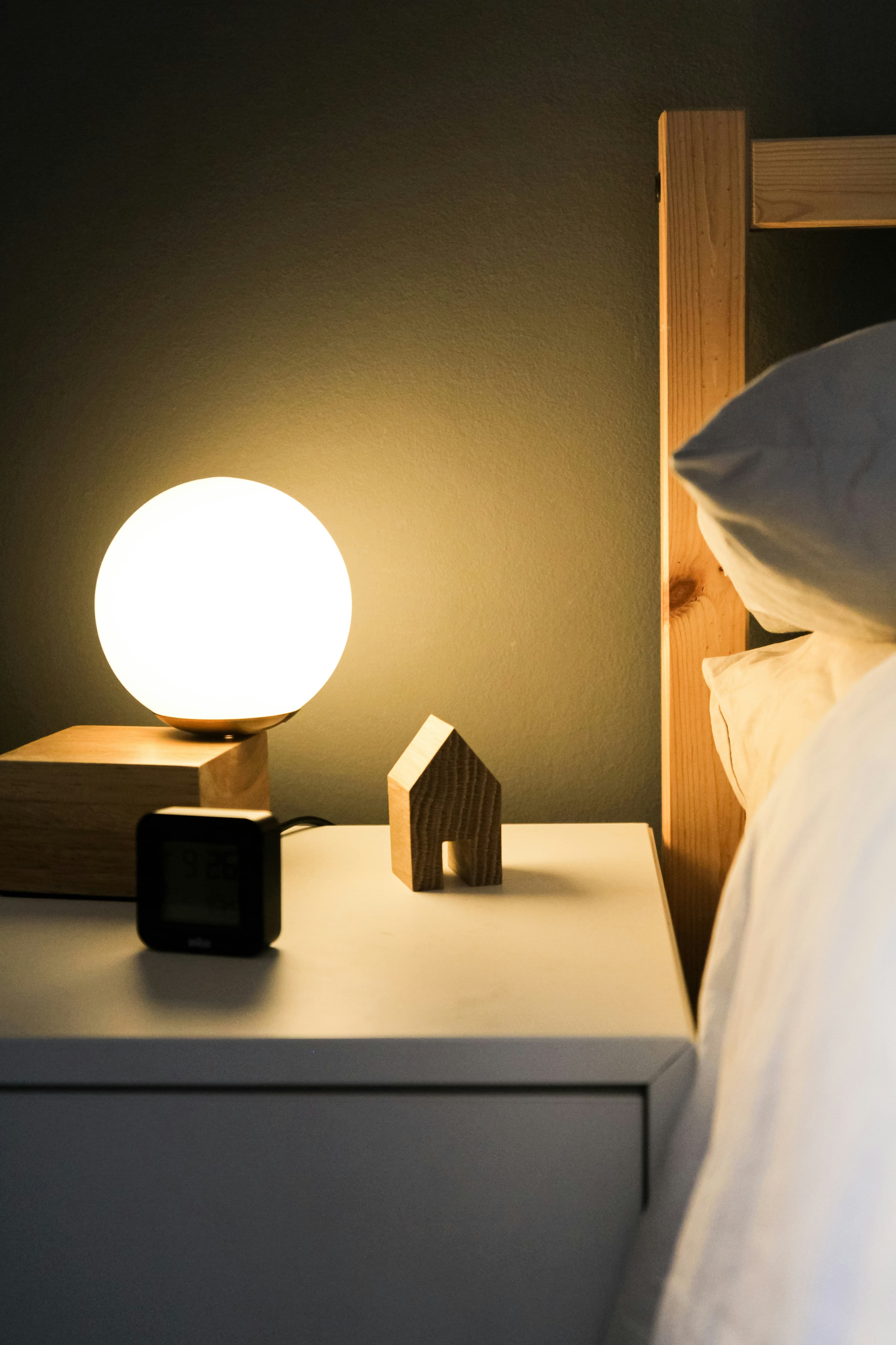 A softly lit bedroom at night. A glowing globe lamp sits on a white nightstand next to a small wooden house figurine and a digital alarm clock. A wooden bed frame and white pillow are visible in the foreground.