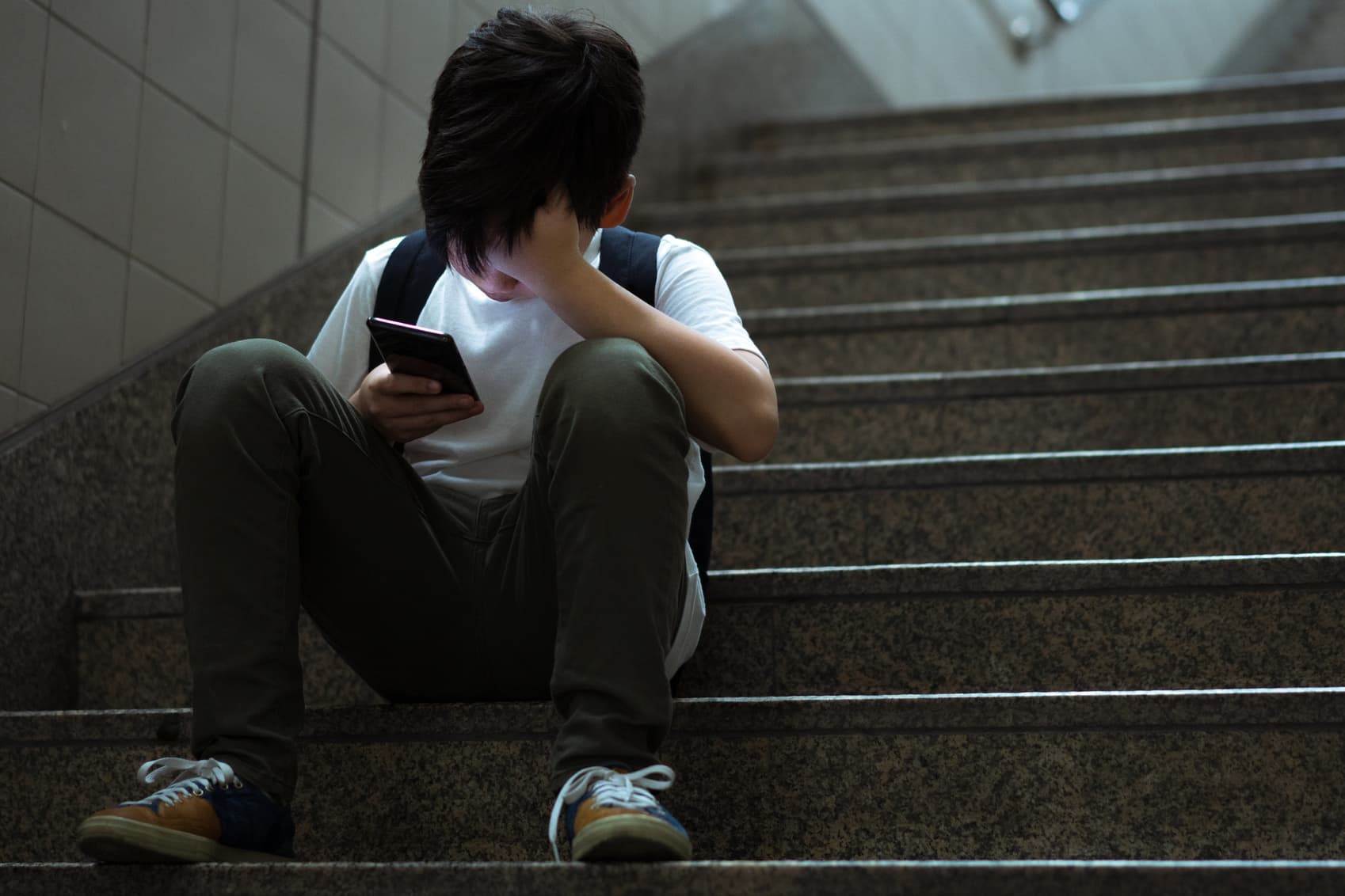 Cyberbullying concept. Young Asian preteen boy sitting at stair, covering his face with hands and other hand holding smartphone.