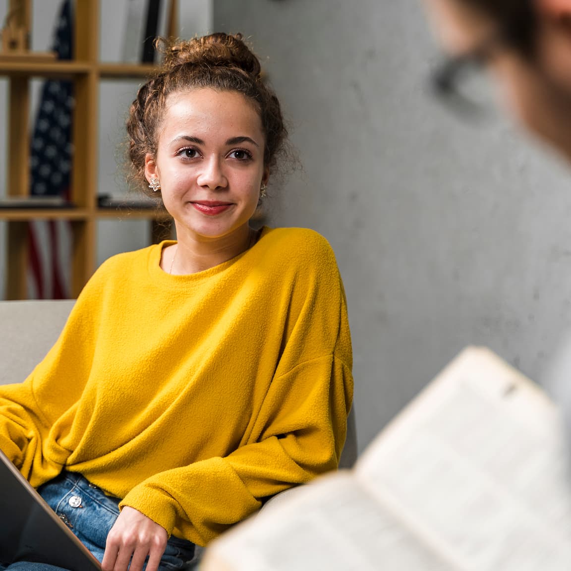 A professional wearing a bright yellow oversized sweater and jeans sits casually during what appears to be an informal meeting. The background shows wooden shelving and an American flag, suggesting an office or workplace setting. Their hair is styled in a casual updo and they're wearing subtle makeup with a warm, engaging expression.